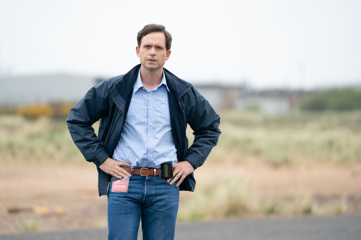 Man standing outdoors in a rural area, wearing a blue jacket, dress shirt, and jeans, hands on hips, with an ID badge and a building blurred in the background. Overcast sky and grassy field add to the open environment.