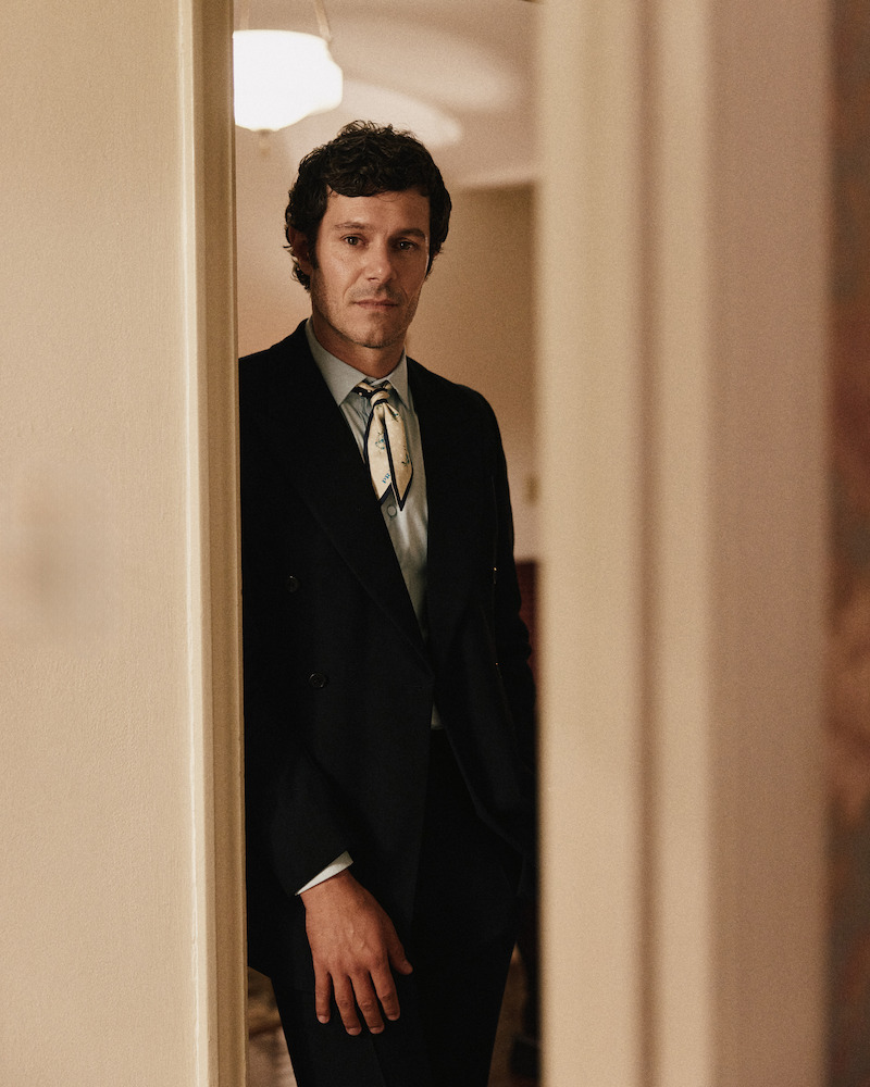 Man in a dark suit with a patterned neck scarf stands in a softly lit, warmly colored indoor hallway, partially framed by a doorway, with a ceiling light in the background.