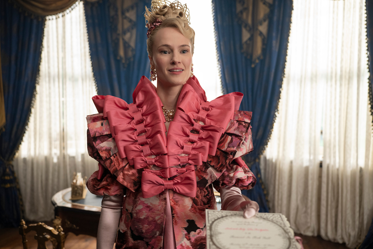 A woman in an elaborate pink and floral Victorian-style dress stands in a luxurious room with blue curtains and ornate furniture, holding a certificate and smiling.