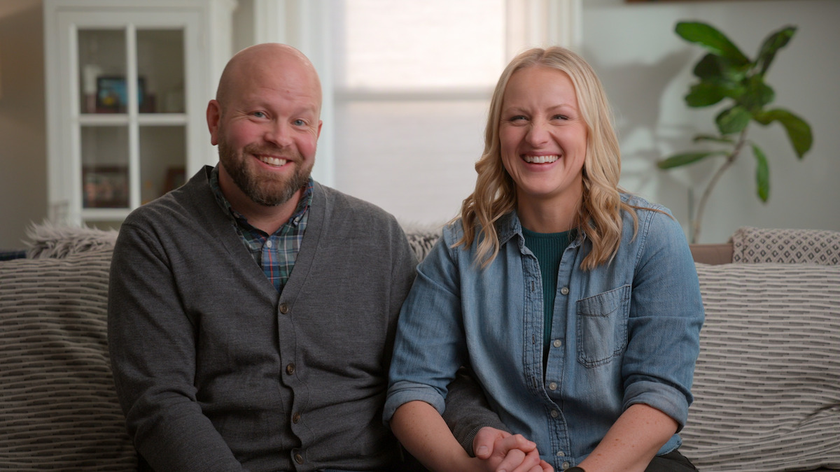 Smiling man and woman sit side by side on a couch in a cozy, well-lit living room with a plant and bookshelf in the background, holding hands and looking at the camera.