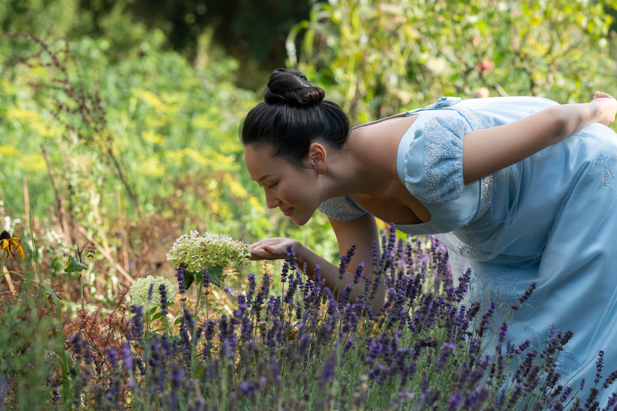 Woman in a light blue historical dress bends to smell flowers in a sunny garden filled with blooming plants and greenery, creating a peaceful and natural outdoor setting.