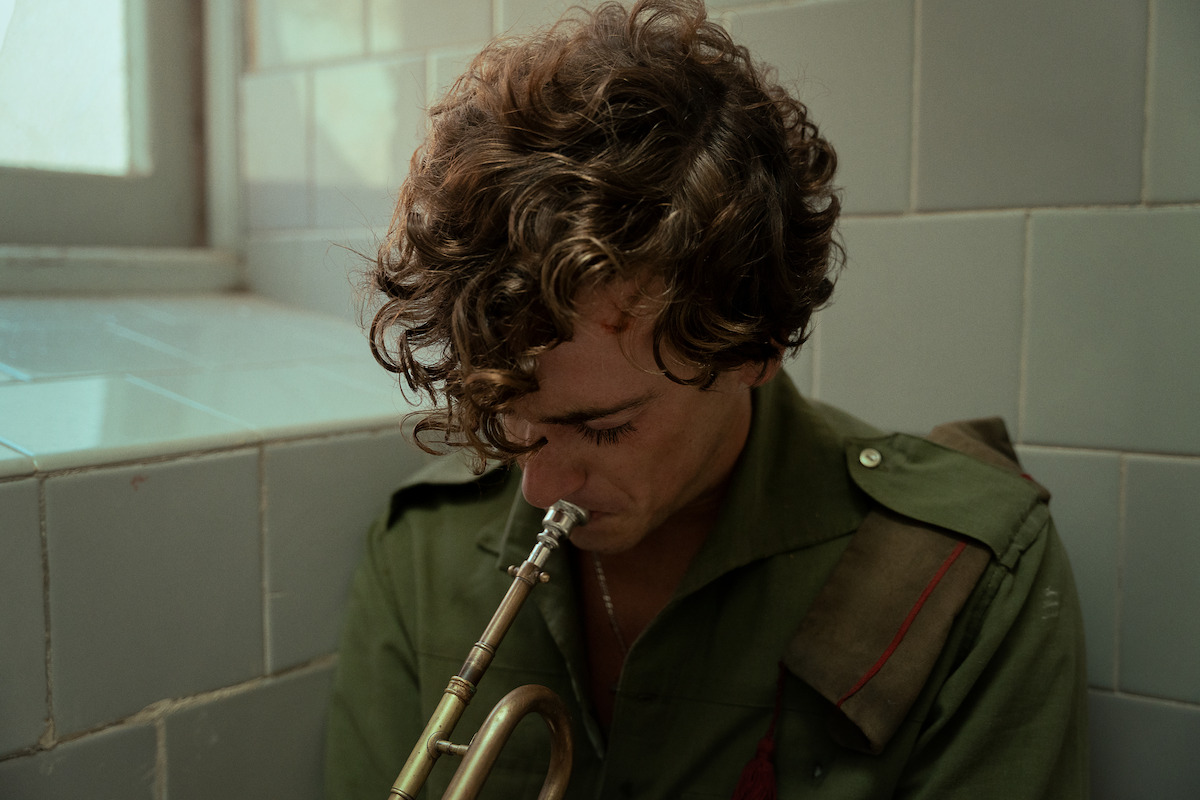 Curly-haired person in military-style green shirt sitting in tiled room by window, looking down while holding a brass trumpet, evoking a somber or reflective mood.