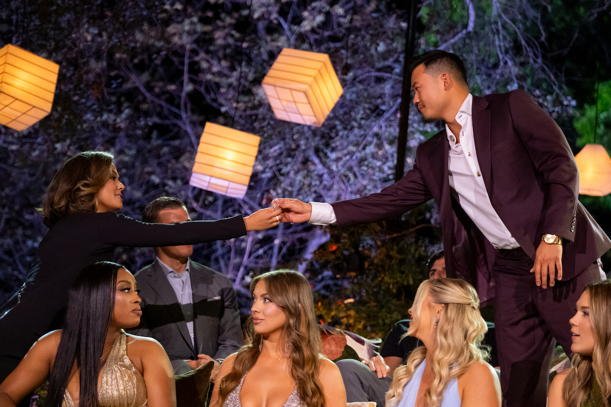 Man in suit reaches to shake hands with seated woman at a formal outdoor gathering, surrounded by other well-dressed people and glowing lanterns, creating a warm, festive, and engaging atmosphere.