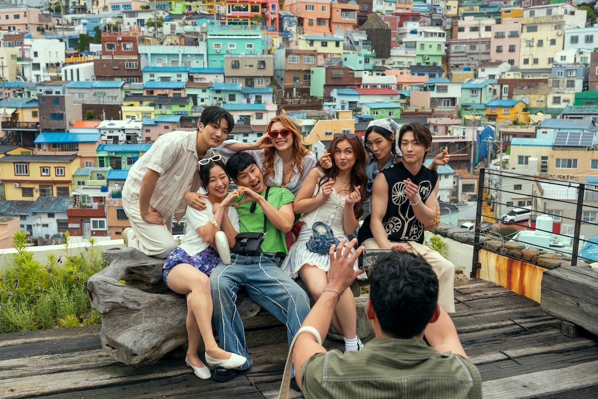 A group of six young adults pose happily on a rooftop with vibrant, colorful hillside houses in the background while another person takes their photo, capturing a lively urban outdoor moment.