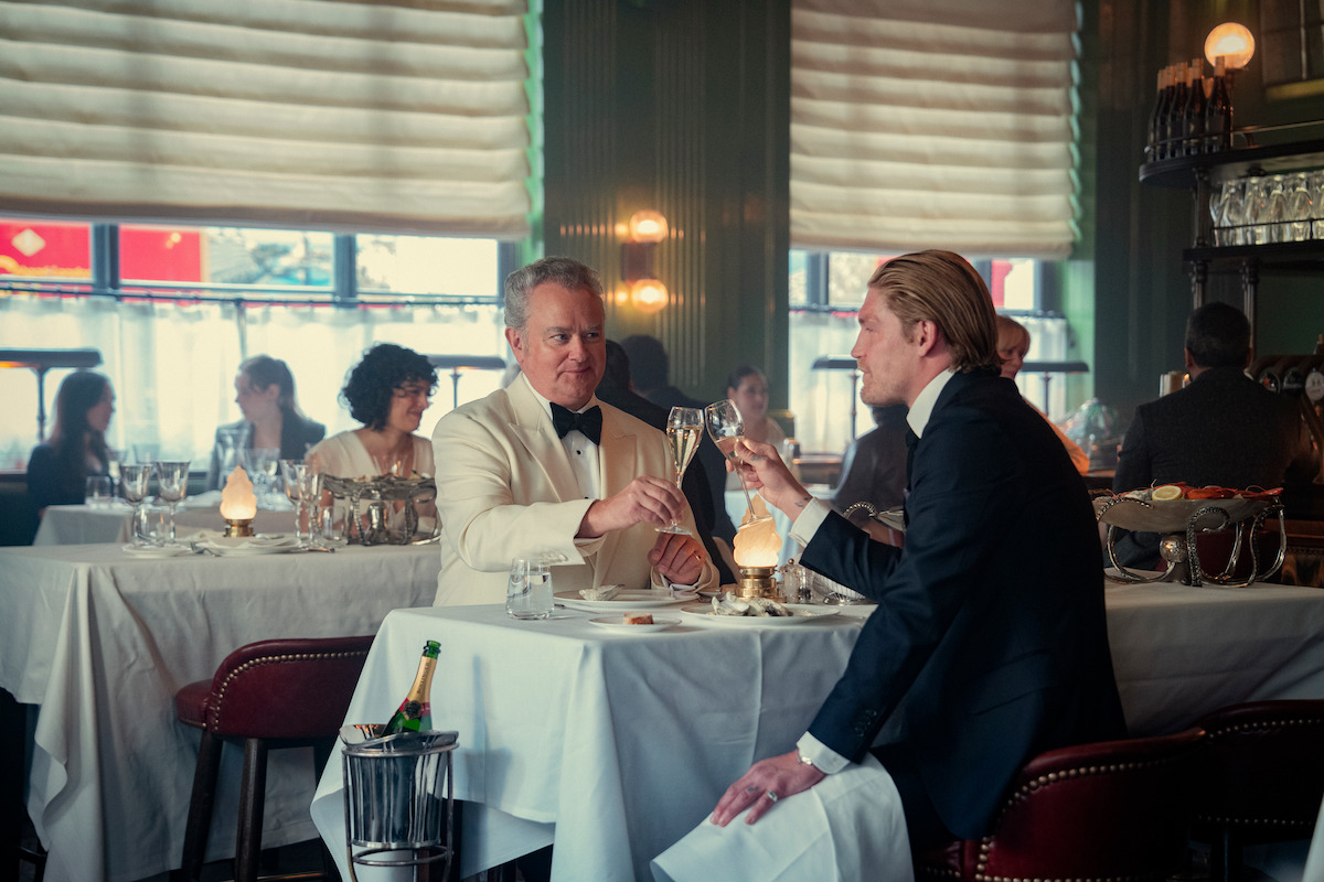 Two men in formal attire toast with wine glasses at a candlelit table in an elegant, upscale restaurant, with other diners in the background, white tablecloths, champagne bucket, and soft, ambient lighting.