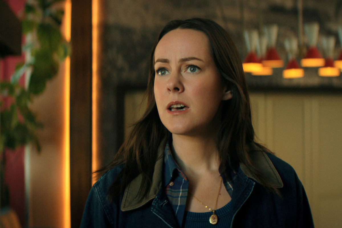 Jena Malone as Claire Cooper looks concerned in a warmly lit indoor setting, wearing a plaid shirt under a denim jacket. Pendant lights and some greenery are visible in the background.