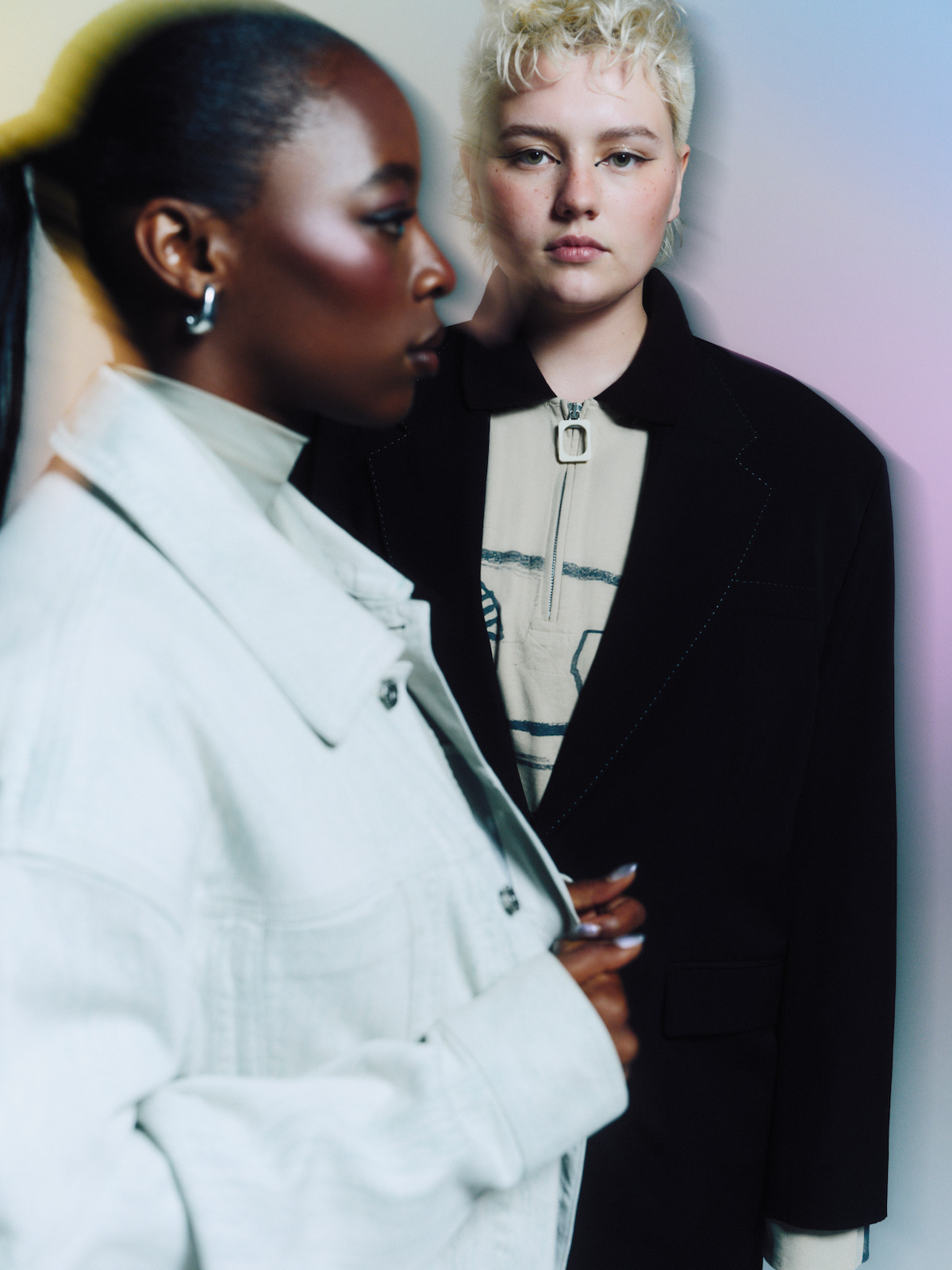 Two people stand indoors against a soft, colorful background. One is in focus wearing a dark blazer, the other is closer to the camera and appears blurred, wearing a light-colored jacket.