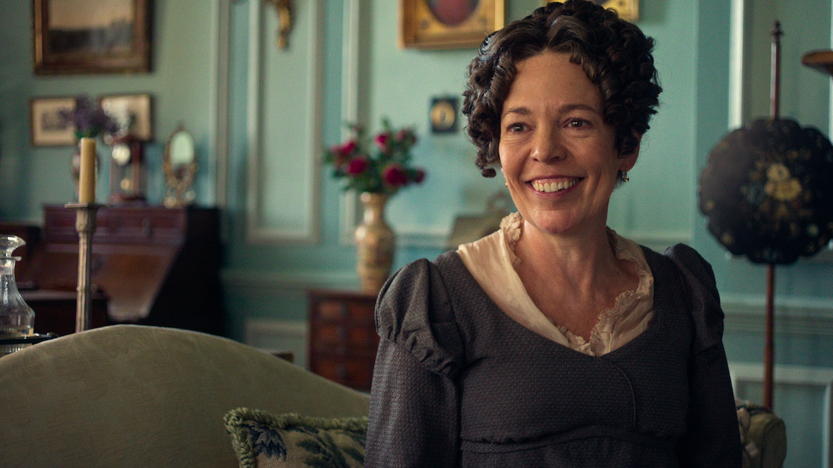 A smiling woman in period costume sits on a couch in a vintage, elegant living room with teal walls, antique furniture, framed art, and a flower vase in the background.