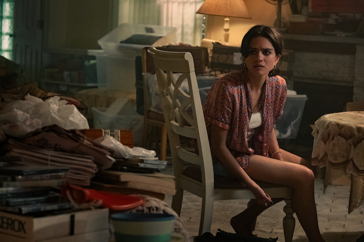 Young woman sitting on a chair in a cluttered, dimly lit room filled with stacks of papers, boxes, and covered furniture, with warm lighting from a table lamp and a window in the background.