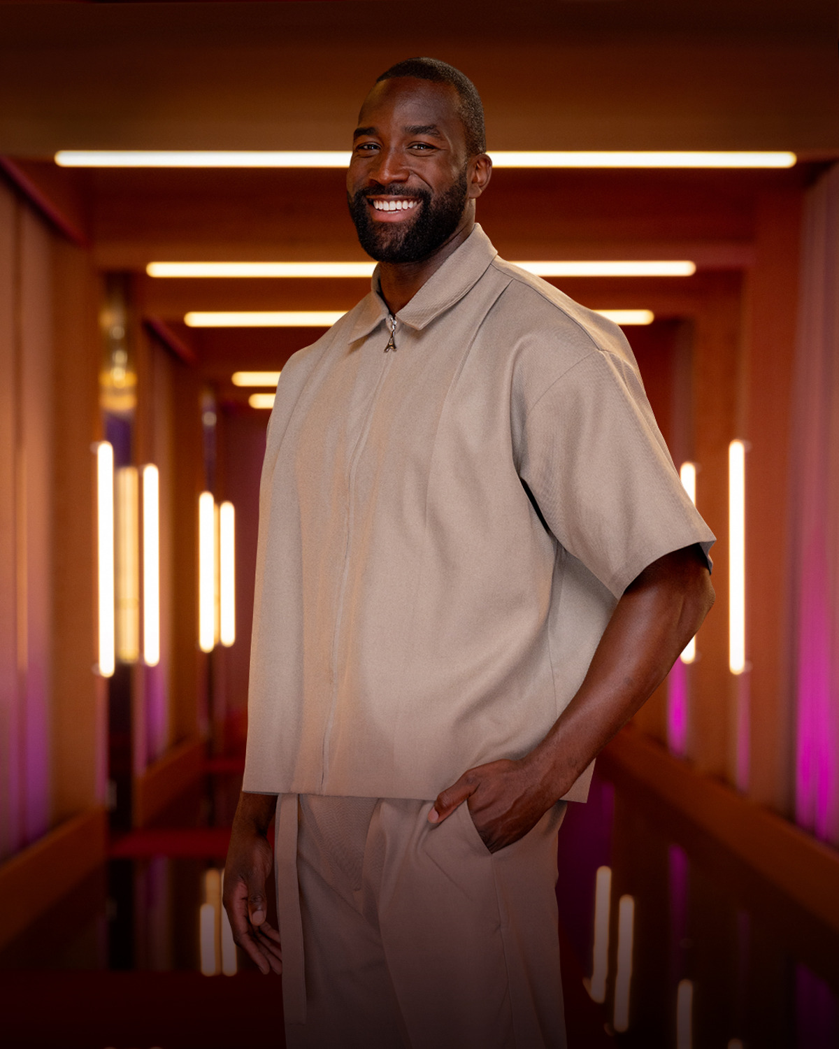 Smiling man wearing a beige shirt and pants stands in a modern hallway with glowing vertical lights and warm, colorful ambiance.