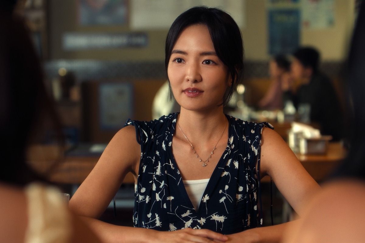 Woman sitting at a table in a restaurant or café having a conversation, with people and tables visible in the softly focused background. Casual, warmly lit social environment.