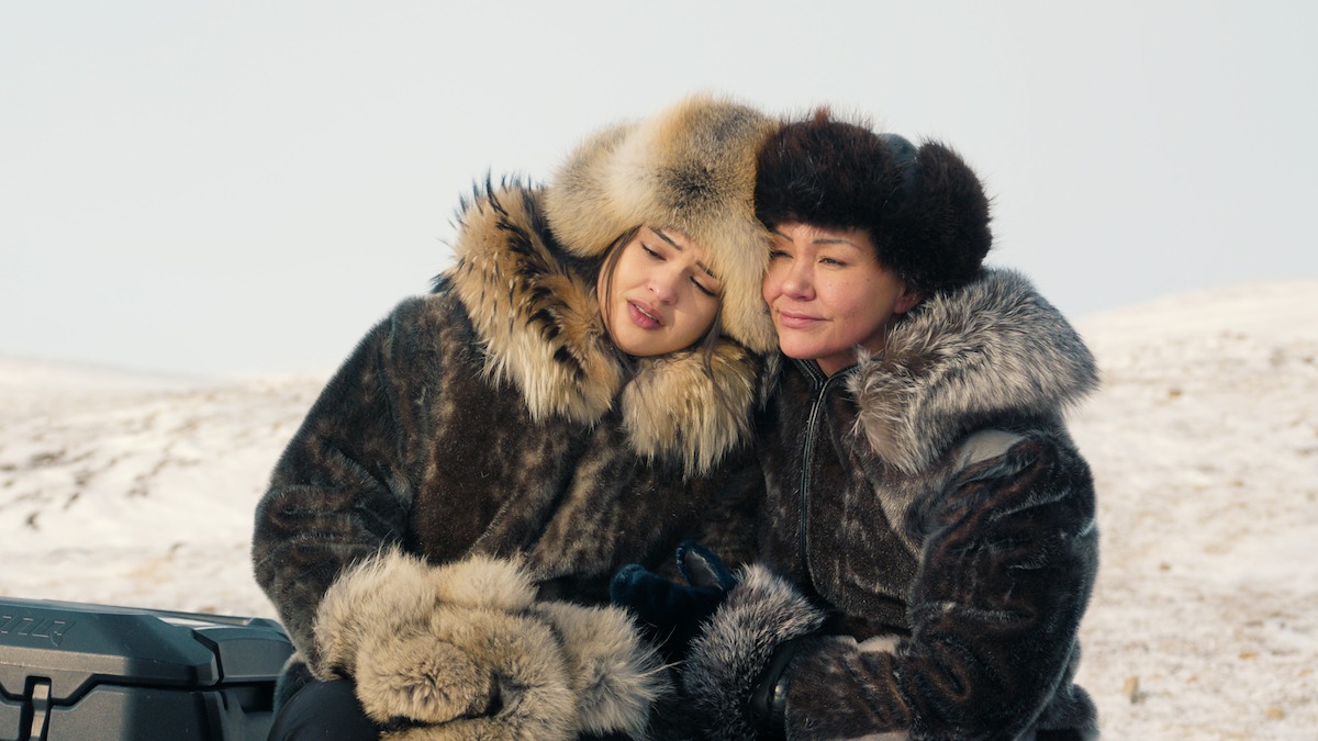Duas pessoas com casacos grossos de pele e gorros estão sentadas bem juntas ao ar livre, cercadas por uma paisagem coberta de neve. Elas compartilham um momento de tranquilidade em meio ao frio.