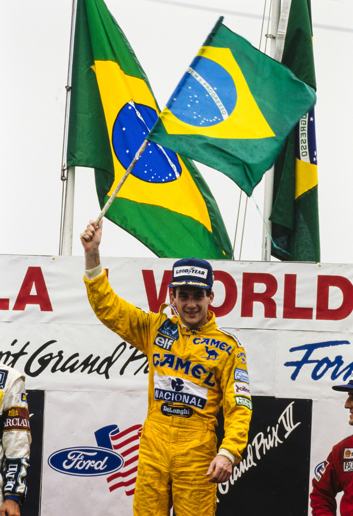 Ayrton Senna comes in first at the Detroit Grand Prix on June 21, 1987, wears a yellow racing jumpsuit, and waves a Brazil flag.