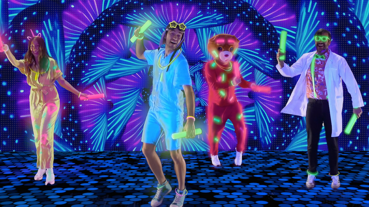 Four people in neon and glow-in-the-dark costumes, one in a bear suit, dance with glow sticks on a vivid, illuminated stage with colorful digital patterns and futuristic lighting effects.