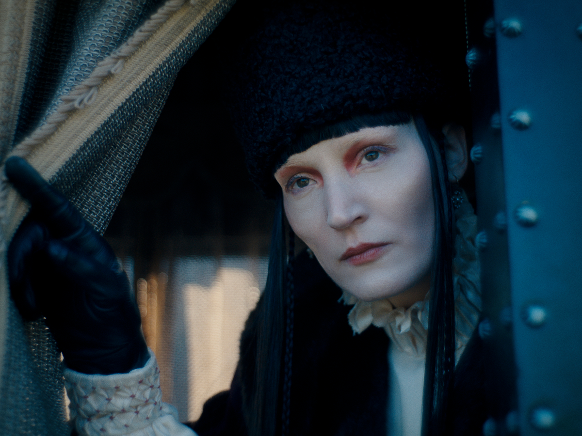 Person with pale makeup and dark clothing, wearing a black fur hat and gloves, peers out from behind ornate curtains in a dimly lit, possibly vintage or historical setting, with soft lighting highlighting their serious expression.