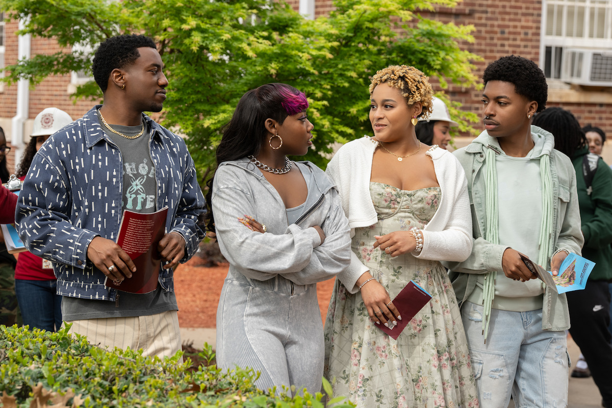 Four young adults stand together outdoors on a college campus, dressed in casual and semi-formal clothes, holding pamphlets and talking, with trees, other people, and a brick building in the background.