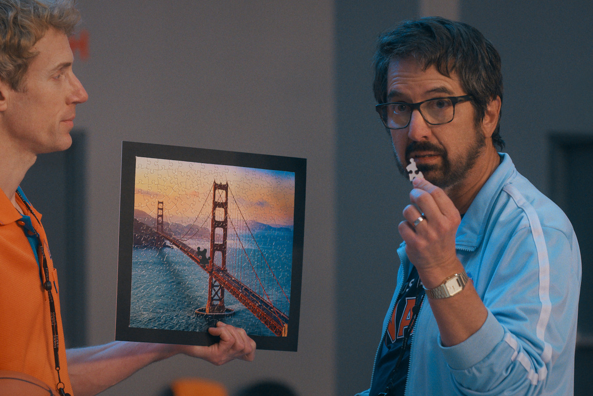 Two men indoors, one holding a finished puzzle of the Golden Gate Bridge at sunset, the other holding a missing piece, both appearing focused in a brightly lit room.