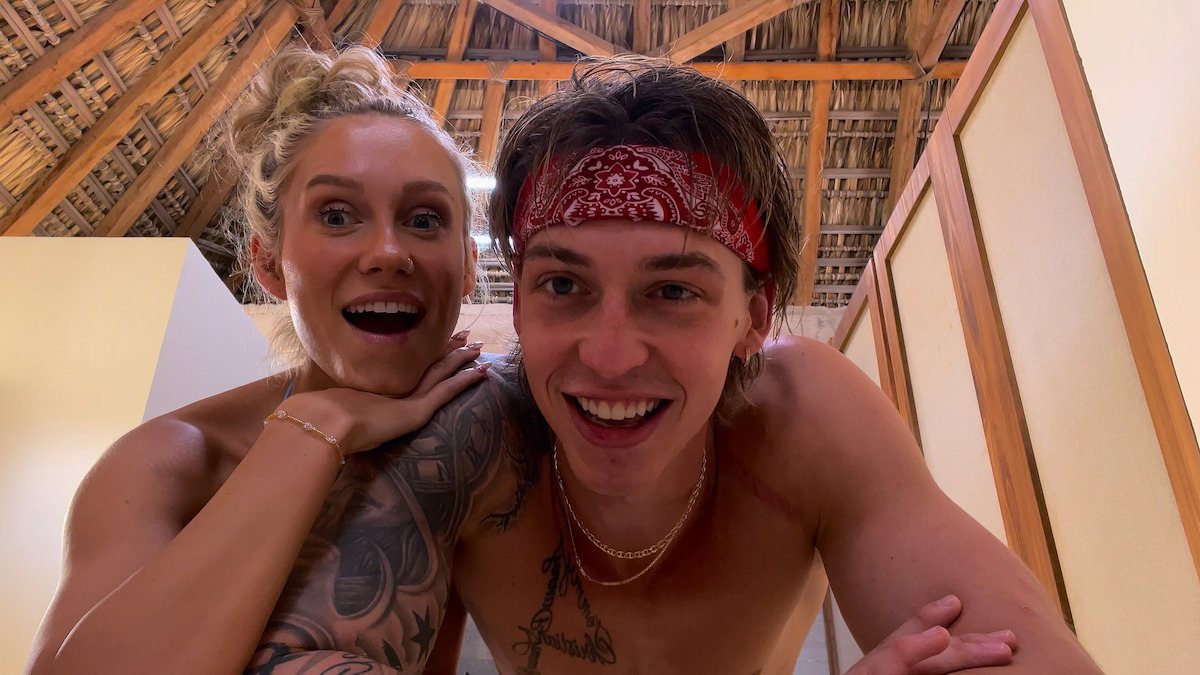 Smiling young man and woman posing together indoors under a rustic wooden ceiling, both shirtless, with the man wearing a red bandana and visible tattoos, appearing happy and relaxed in a casual, tropical setting.