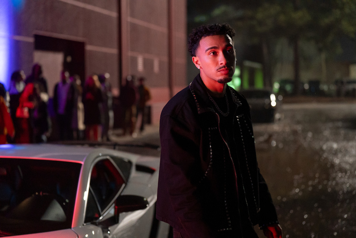 A young man stands beside a silver car at night, illuminated by colorful lights. A line of people waits by a building in the background, creating a moody, urban nightlife atmosphere.