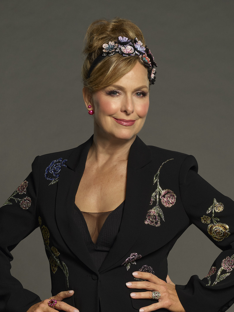 Woman in a black floral-embroidered blazer and bejeweled headband poses confidently with hands on hips, wearing pink earrings and rings, in a studio with a plain, neutral background.
