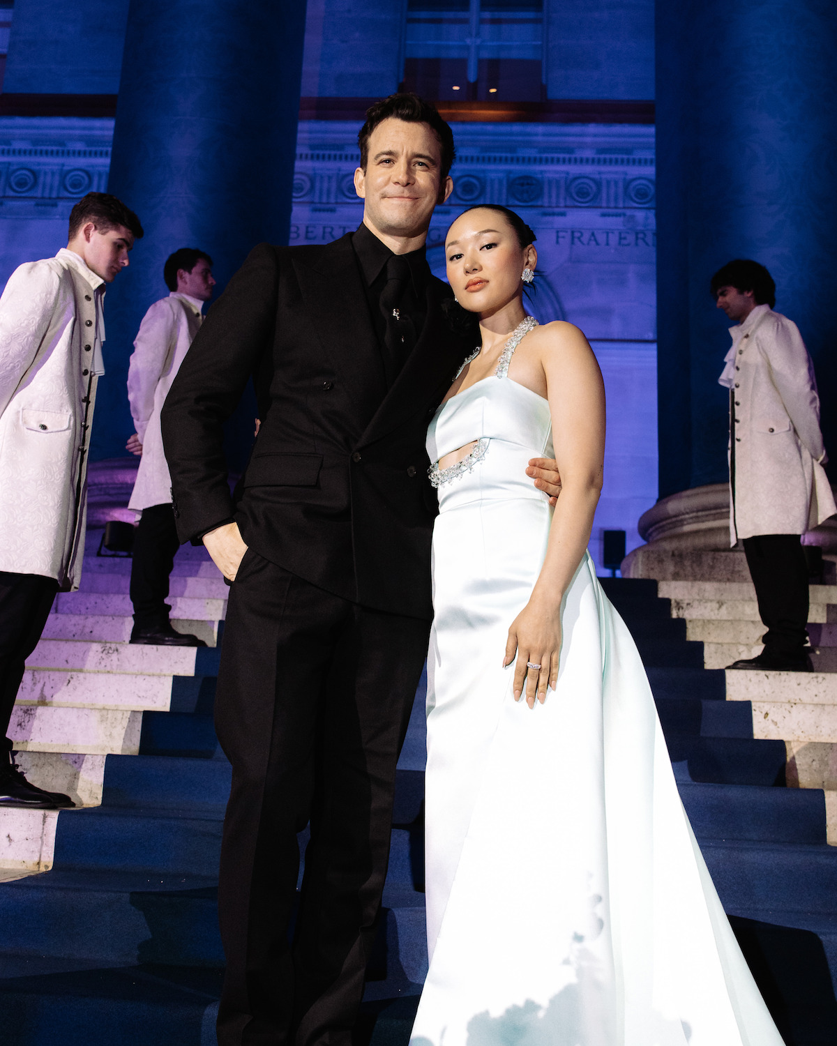 A man in a black suit and a woman in a white gown pose together on blue-lit steps at night, with four people in light-colored coats standing on the stairs behind them in front of a grand building with columns.