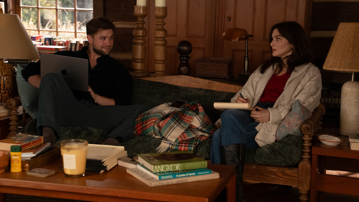 Leo Woodall as Vladimir and Rachel Weisz as The Protagonist sitting on a cozy couch in a wood-paneled cabin, surrounded by books, a candle, and a plaid blanket, having a conversation. One holds a laptop, the other a notepad.Leo Woodall as Vladimir and Rachel Weisz as The Protagonist sitting on a cozy couch in a wood-paneled cabin, surrounded by books, a candle, and a plaid blanket, having a conversation. One holds a laptop, the other a notepad.