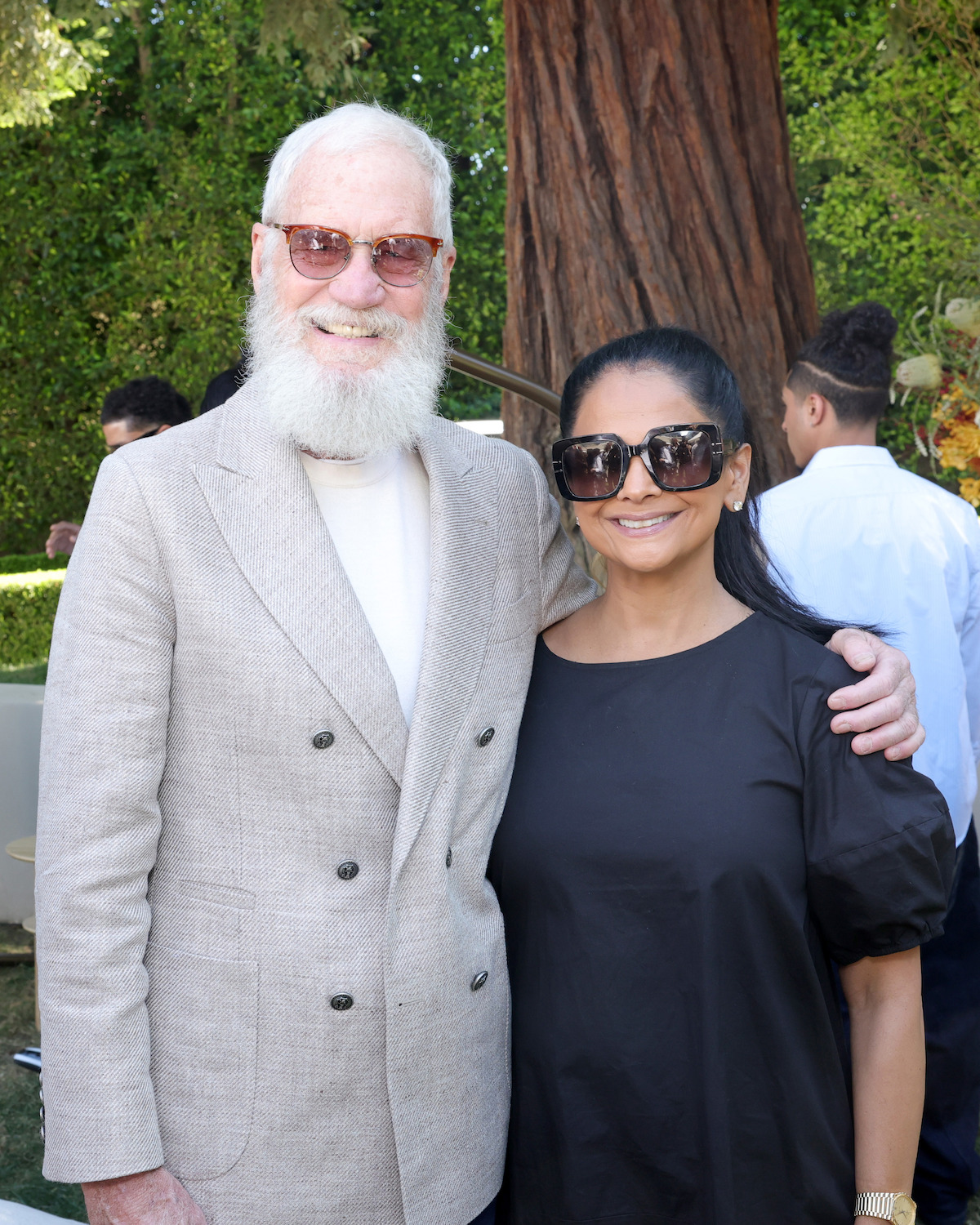 Two people smiling outdoors at a daytime event, standing in front of a large tree with greenery in the background. Both are wearing sunglasses and casual elegant clothes, with other people and decorations visible behind them.