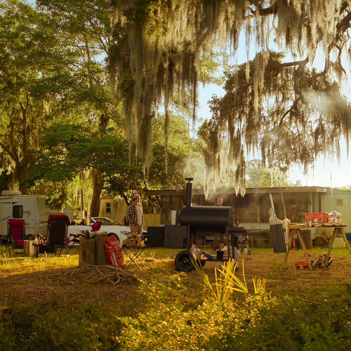 Uma pessoa está em pé diante de uma churrasqueira enfumaçada em ambiente ao ar livre com cadeiras, mesas, equipamentos de camping e trailers debaixo de árvores cheias de musgo espanhol, em um clima tranquilo e rústico.