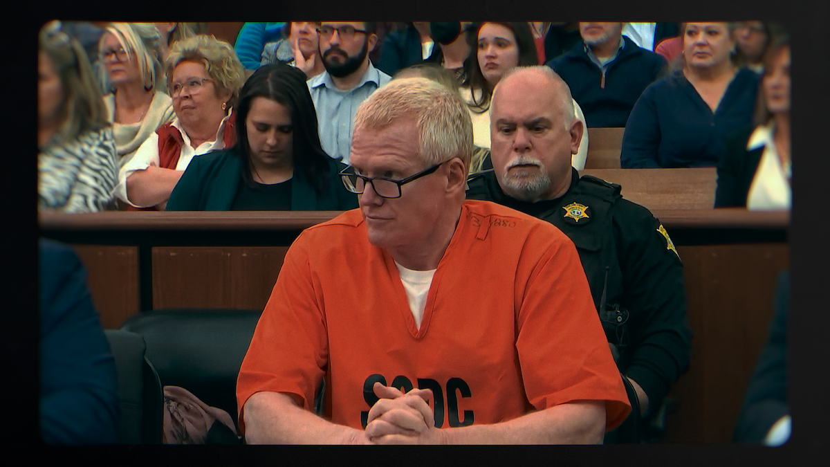 Man in orange prison uniform sitting in a courtroom, hands clasped, with a guard behind him and an audience of people seated in the background, some watching attentively and others looking down.