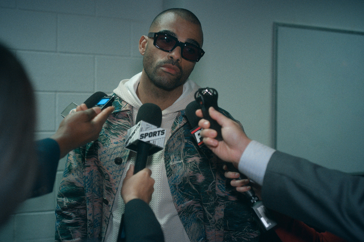 A man wearing sunglasses and a patterned jacket is standing against a white brick wall while multiple reporters hold microphones toward him, suggesting a press interview or media event in an indoor setting.