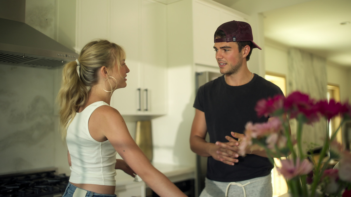 Two young adults talking in a modern kitchen, one wearing a white tank top and earbuds, the other in a black shirt and backwards cap. Bright, natural light fills the space, and pink flowers are blurred in the foreground.