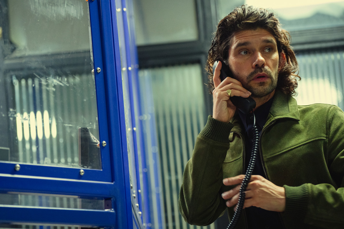Homem com cabelo longo e cacheado, usando uma jaqueta verde, fala ao telefone em uma cabine telefônica azul, com expressão alerta ou preocupada, em um ambiente urbano ao fundo.