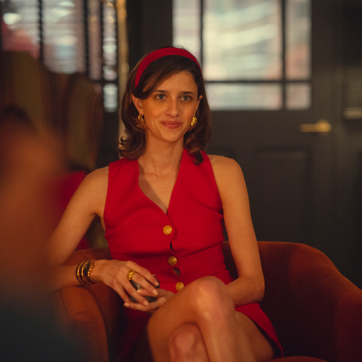Woman in a sleeveless red dress with gold buttons and matching headband sits on an orange chair in a warmly lit, stylish indoor setting with blurred background and glass doors.