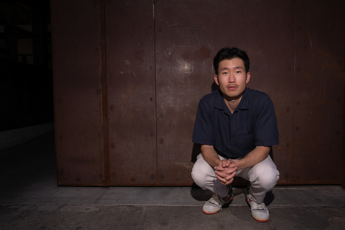 Man squatting on concrete in front of a large rusty metal wall at night, wearing a navy shirt, white pants, and sneakers, looking directly at the camera in an urban outdoor setting.