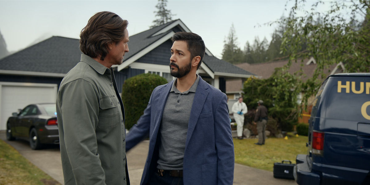 Two men talking seriously outside a suburban house with forensic investigators working in the background and a law enforcement van parked nearby, suggesting an active crime scene investigation.