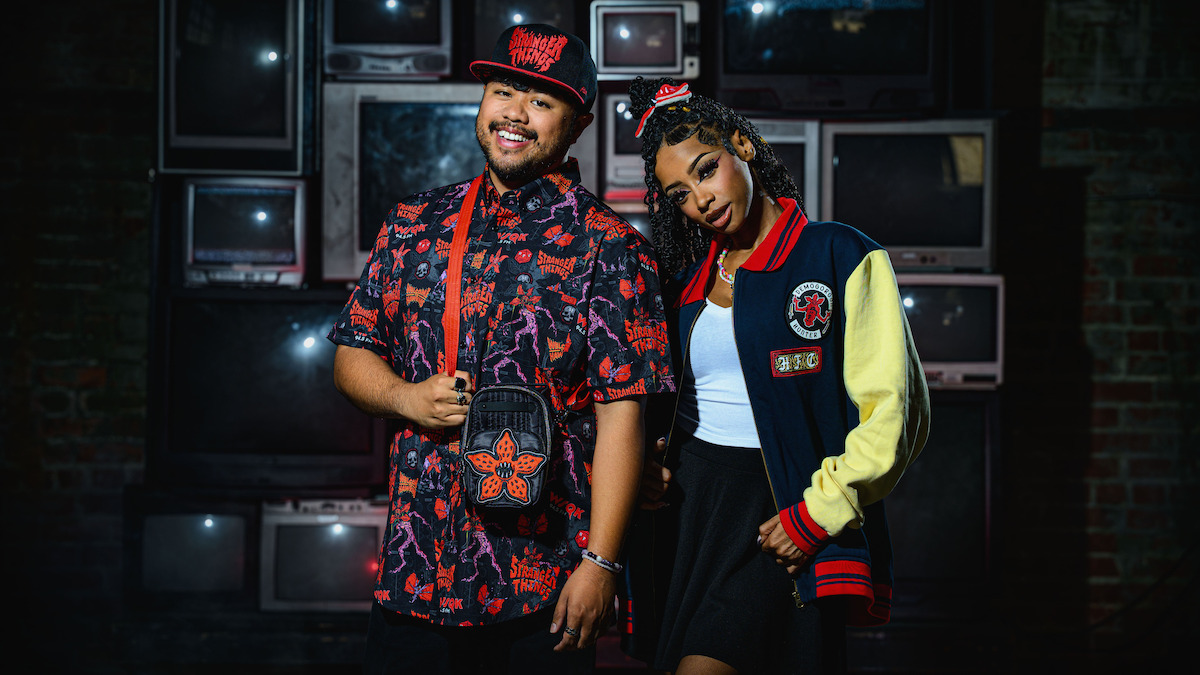 Two stylish young adults, one in a patterned shirt and cap, the other in a varsity jacket, stand smiling in front of a wall of vintage CRT televisions in a dimly lit urban setting.