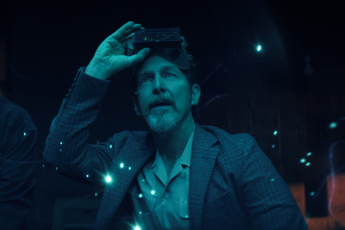 Denis O’Hare as Wally, in a suit holding a futuristic headset on his head sits in a dimly lit, high-tech environment with glowing blue lights and floating particles surrounding him, suggesting a science fiction or virtual reality setting.