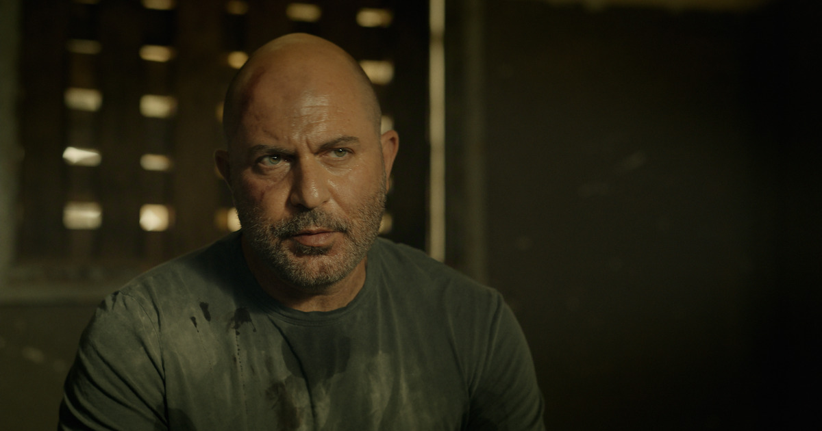 Bald man with a beard sitting indoors in dim lighting, wearing a dark shirt with stains. The background shows a wall with wooden slats and a soft light entering the room, creating a moody, tense atmosphere.