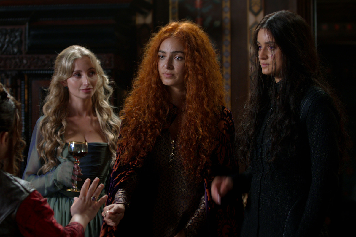 Three women in medieval fantasy clothing stand indoors, one holding a goblet, engaged in conversation with a seated figure. The setting is ornate, with dark wood and intricate decor, creating a mysterious and serious atmosphere.
