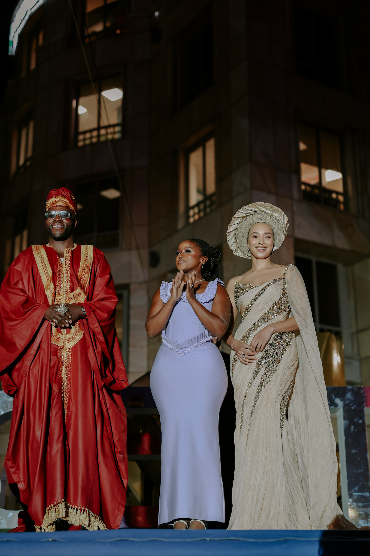 Three people in elegant attire stand on a stage outdoors at night, with a tall building and illuminated windows in the background.