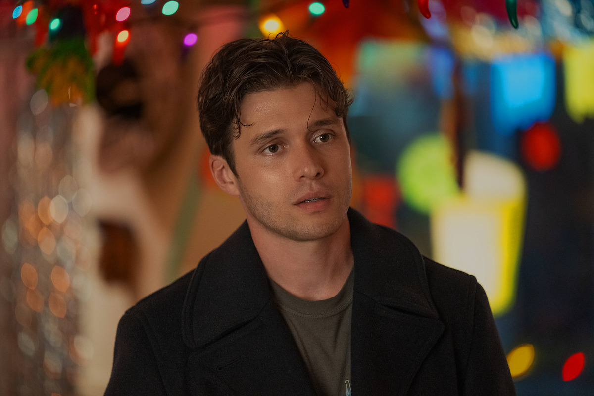 Young man with brown hair in a dark coat stands indoors beneath colorful string lights, with a blurred vibrant background of decorations and soft lighting.