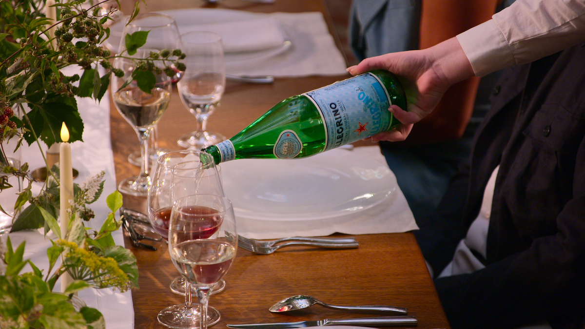 An image featuring a table with someone’s arm reaching through two seated people to pour S.Pellegrino from a bottle into a wine glass.