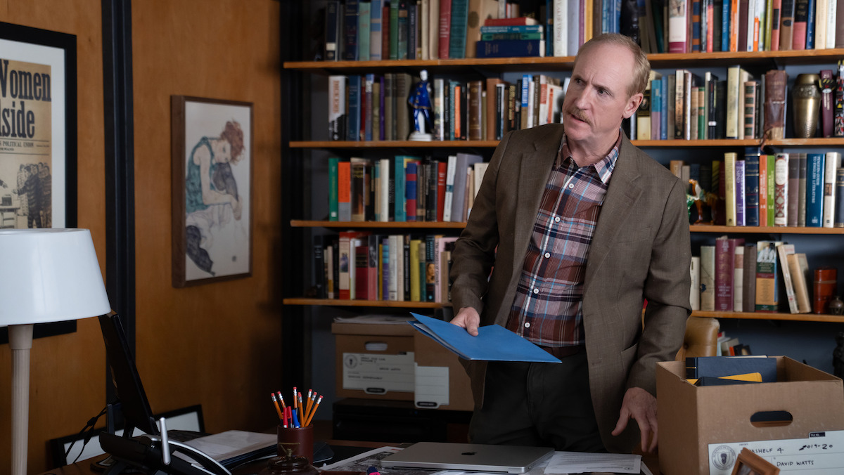 A man in a brown blazer stands in a cluttered office, holding a blue folder. He's surrounded by boxes, bookshelves filled with books, a desk with office supplies, and framed art on the wall, suggesting an academic or professional setting.
