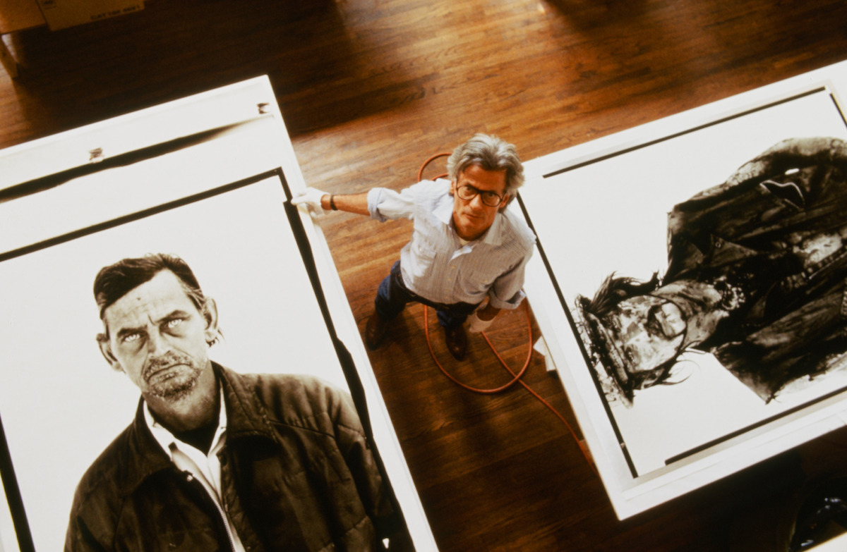 An artist stands in a spacious studio with wooden floors, surrounded by large black-and-white portrait artworks displayed on tables, looking up at the camera from above.