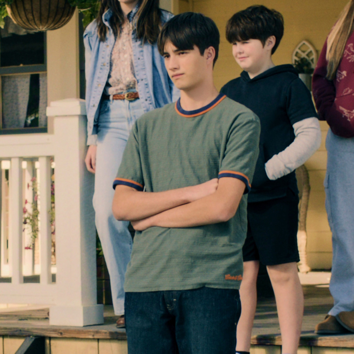 A group of kids stands on the front porch of a house. The boy in front crosses his arms, while others stand or walk behind him. The scene is outdoors, in a suburban residential setting with plants and wooden steps.