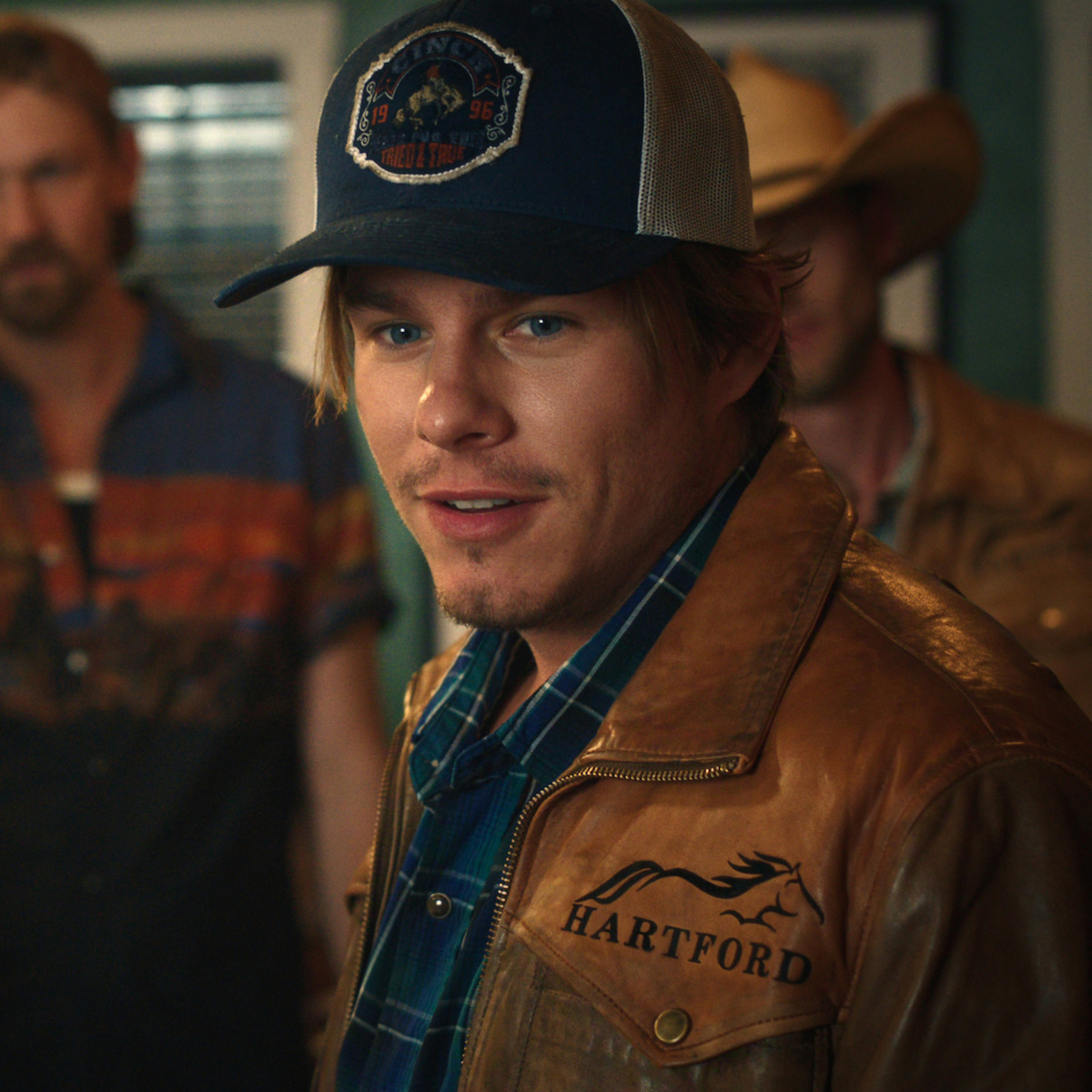 Young man in brown leather Hartford jacket and blue plaid shirt, wearing a trucker hat, stands indoors with two men in Western attire in the background, creating a casual, country setting.