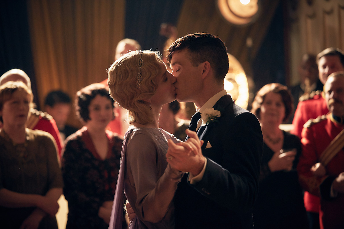 A couple in formal attire shares a kiss while dancing at a vintage-themed party or wedding, surrounded by elegantly dressed guests in a warmly lit, classic ballroom setting.