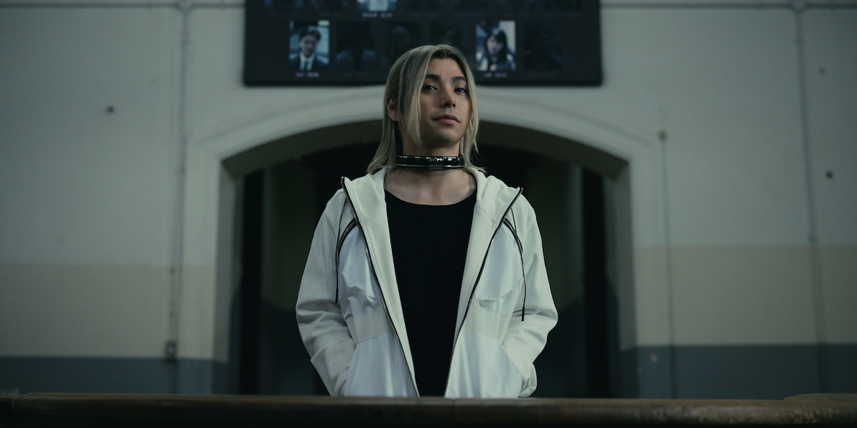 Person with long blond hair and white jacket stands indoors in front of an arched doorway and a wall with photos. Industrial setting with muted colors, the individual appears to be wearing a metallic collar or choker around the neck.
