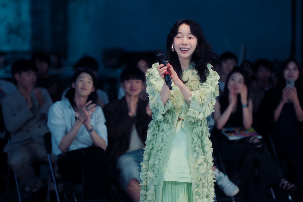 A woman in a green outfit stands smiling and holding a microphone, speaking to an audience seated in a dimly lit room, with several people clapping in the background.