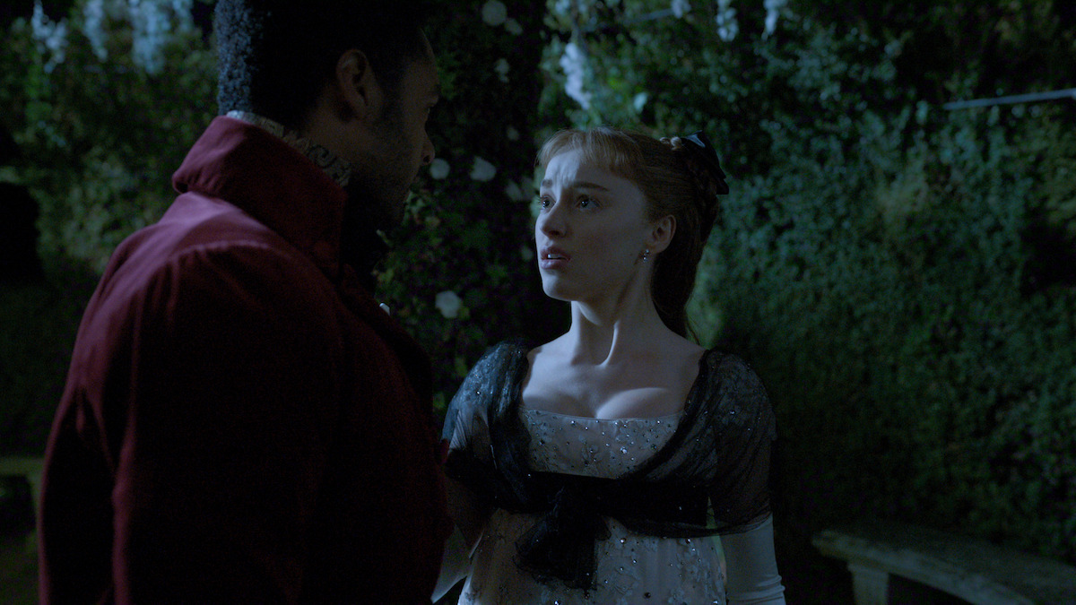 A man and woman in period clothing stand close together in a garden at night, their expressions intense, with greenery and flowers visible in the dimly lit background.